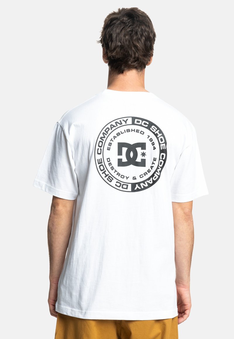 Bílý bavlněný tričko s černým kulatým logem na zádech. Obsahuje text: "DC SHOE COMPANY" a "ESTABLISHED 1994." Krátké rukávy.