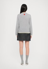 Grauer, gestrickter Pullover mit geripptem Saum und Bündchen, kombiniert mit einem dunklen Denimsrock. Auffälliges rotes Etikett auf der Rückseite, graue knielange Socken und Absätze.