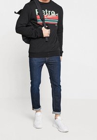 Schwarzer Pullover mit "Retro"-Schriftzug und bunten Streifen, kombiniert mit dunkelblauen Jeans und weißen Turnschuhen. Legeres Outfit mit Rucksack.