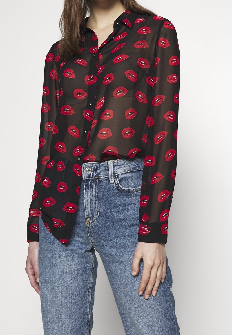 Sort gennemsigtig bluse med et mønster af røde læber, lange ærmer, knaplukning foran og kontrasterende sorte manchetter; båret over lyseblå jeans.