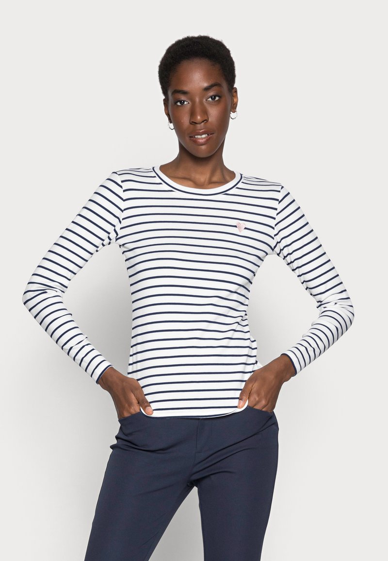 Chemise à manches longues avec des rayures horizontales blanches et bleu marine, col rond et petit emblème en forme de cœur sur la poitrine. Tissu lisse, design ajusté.