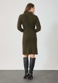 Olíva zöld kötött ruha hosszú ujjakkal, testhez simuló derékkal és egyenes sziluettel, magas fekete bőr sarkú csizmával párosítva.