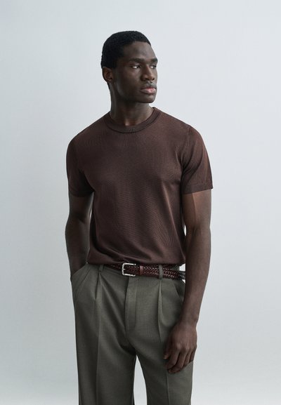 Homme portant une chemise à manches courtes marron foncé et un pantalon plissé gris avec une ceinture tissée, debout les mains dans les poches, regardant sur le côté.