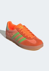 Oranžové a růžové semišové tenisky se zelenými pruhy, gumovou podrážkou a texturovaným jazykem. Obsahují branding na jazyku a patě.