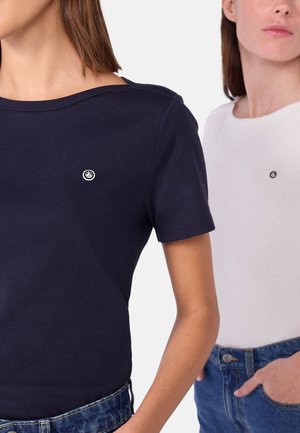Due donne che indossano t-shirt a manica corta con scollatura a girocollo, una blu navy e l'altra bianca, abbinate a jeans blu, che si voltano leggermente lontano dalla macchina fotografica.