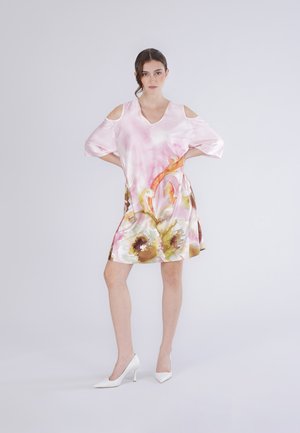 Femme portant une robe rose à fleurs avec épaules dénudées et des talons blancs, debout les mains sur les hanches devant un fond clair uni.