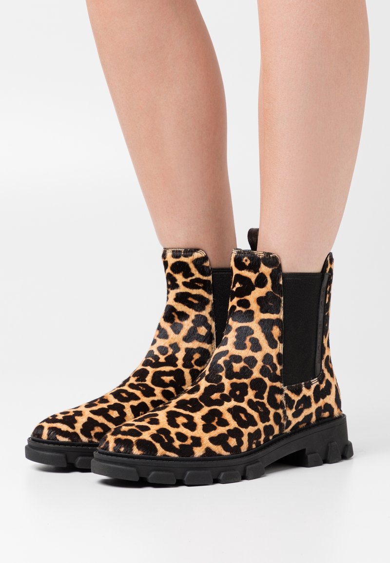 Bas de jambes montrant des bottes longueur mollet à imprimé léopard avec des côtés en élasthanne noir et des semelles épaisses en caoutchouc sur un fond uni.