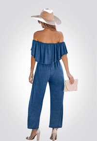 Navy jumpsuit zonder schouders met een gerimpelde top, brede benen en een textuurstof. Het model houdt een beige clutch vast en draagt een strohoed en hakken.