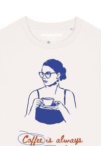 Witte t-shirt met een blauwe illustratie van een vrouw die een theekopje vasthoudt, met daaronder de tekst "Koffie is altijd" in oranje.