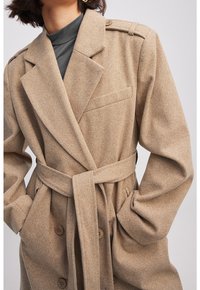 Beige visgraatjas met brede revers, een ceintuur in de taille, een enkele borstzak en grote bruine knopen aan de voorkant.
