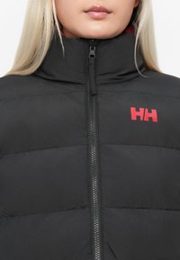 Veste matelassée noire avec col montant, fermeture éclair à l'avant et logo rouge en accent. Présente une texture lisse et mate avec des sections matelassées.