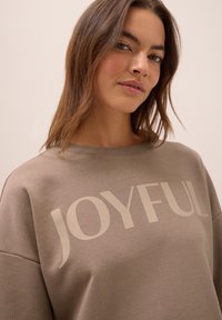 Ung kvinna med ljust brunt hår bär en taupe sweatshirt med stor "JOYFUL"-text över bröstet, neutral bakgrund.