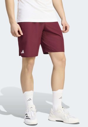 Person trägt bordeauxrote Sportshorts, weiße Sportschuhe und weiße kniehohe Socken mit sichtbarem Adidas-Logo auf Socken und Shorts.