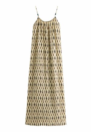 Kobieta ubrana w bezrękawową beżową sukienkę maxi z czarnymi wzorami w kształcie diamentów, stojąca boso na puszystym dywanie w słonecznym salonie.