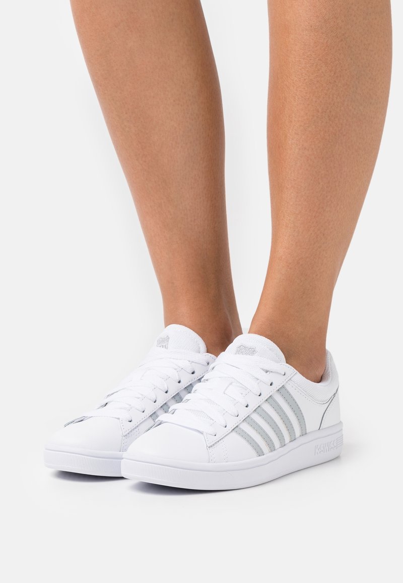 Weiße Sneakers mit drei silbernen Streifen, aus glattem synthetischen Material. Verfügt über weiße Schnürsenkel und eine strukturierte Gummisohle.
