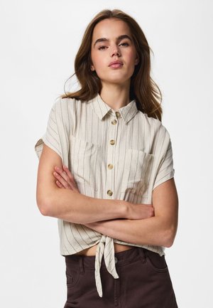 Femme portant une chemise beige à rayures à manches courtes, boutonnée et nouée à la taille, ainsi qu'un pantalon foncé, debout, bras croisés devant un fond uni.
