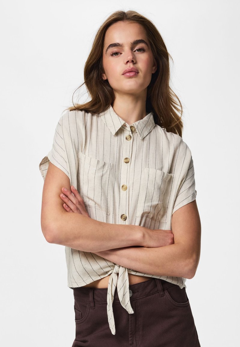 Donna che indossa una camicia beige a righe con maniche corte, allacciata in vita, e pantaloni scuri, in piedi con le braccia incrociate davanti a uno sfondo semplice.