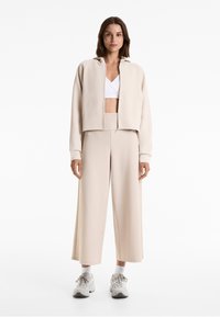 Beige geribbelde zip-up jack met capuchon, gecombineerd met bijpassende wijde broek. Draagover een witte cropped top, en afgewerkt met lichtgekleurde sneakers.