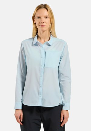Femme portant une chemise bleu clair à manches longues avec des boutons et une poche poitrine, debout devant un fond blanc uni.