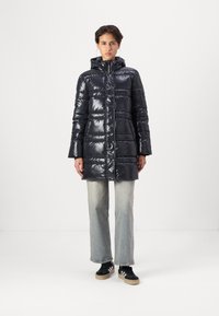 Svart pufferjakke med en blank finish, glidelås foran, hette og horisontalt quiltet mønster. Kombinert med lyseblå utsvingte jeans og svarte joggesko.