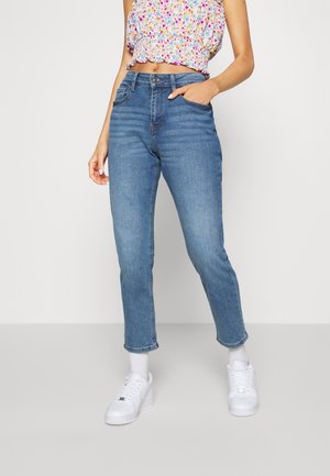 Vrouw draagt blauwe jeans met hoge taille, witte sneakers en een cropped bloemen top, staat met één hand in haar zak.