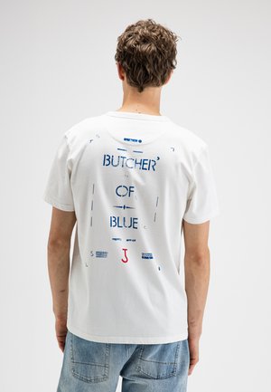 Mann mit lockigem Haar, der ein weißes T-Shirt mit blauem und rotem Text „BUTCHER' OF BLUE“ auf dem Rücken und hellblaue Jeans trägt, zeigt sich von hinten.