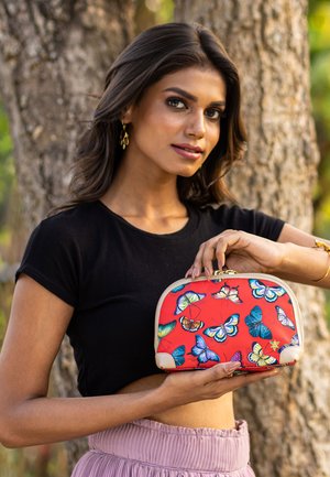 Vrouw in een zwart topje en een roze rok die een rode tas met kleurrijke vlinderprints vasthoudt, staand voor een boom buiten.