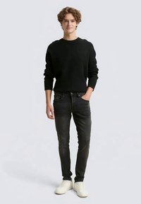 Jovem em pé com uma mão no bolso, vestindo um suéter preto, jeans pretos e ténis brancos sobre um fundo claro.