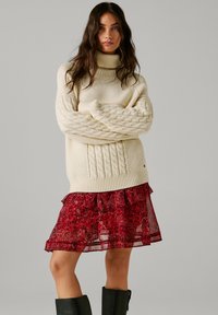 Paksu kerros neulottu villapaita, jossa on korkea kaulus ja teksturoidut hihat, kerrostettuna ohuen, röyhelöisen punaisen kukkamekon päälle. Mustat polvimittaiset saappaat.