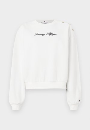 Fehér, rövidre szabott pulóver, puha anyagból készült, hosszú ujjakkal, bordás mandzsettával, és egy fekete hímzett logóval az elején. Egyik vállán három gomb díszítés.