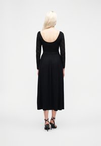 Γυναίκα με μακριά ξανθά μαλλιά, που φοράει μαύρο δαντελένιο τοπ με μακριά μα νίκια και μαύρη μίντι φούστα, στέκεται με την πλάτη προς τα εμπρός, φορώντας μαύρα σανδάλια με ψηλό τακούνι.