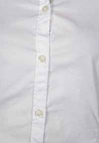 Camicia bianca in cotone con chiusura a bottoni, dotata di bottoni in plastica, cuciture visibili e una texture liscia, leggermente stropicciata.