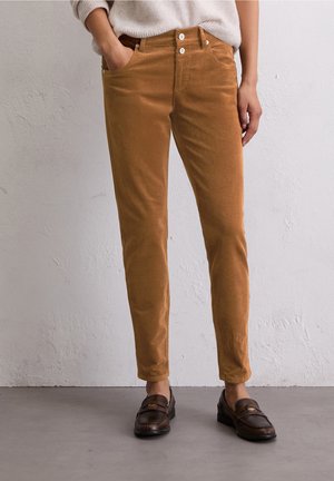 Persona vistiendo pantalones de pana marrón ajustados con cierre de doble botón y mocasines marrón oscuro, de pie contra una pared clara con textura.