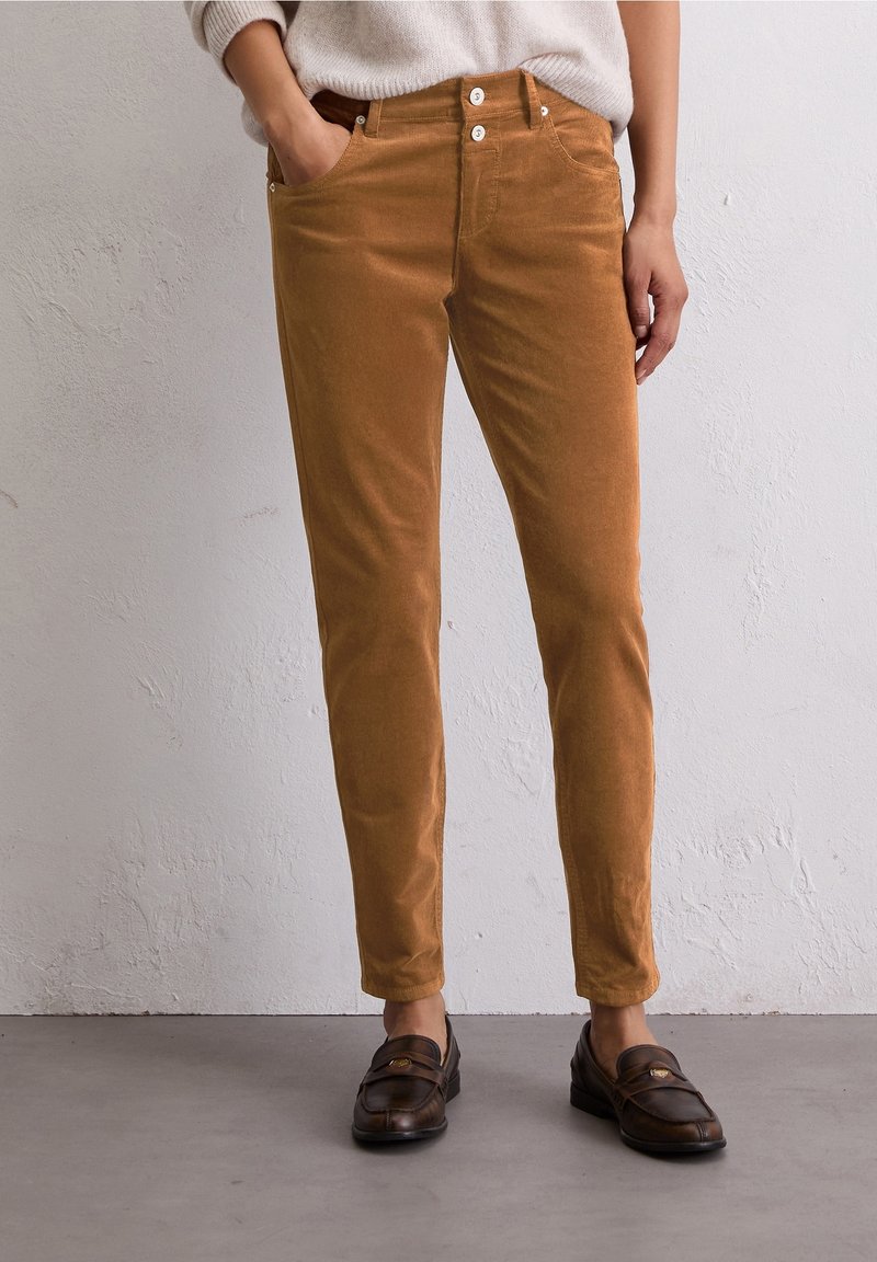 Personne portant un pantalon en velours côtelé marron coupe slim avec fermeture à double bouton et des mocassins marron foncé, debout contre un mur texturé clair.