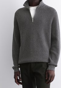 Pull gris côtelé avec un col montant, doté d'une fermeture éclair demi-zip. Fabriqué en tissu tricot texturé, associé à un pantalon vert foncé.