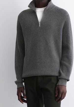 Grauer gerippter Pullover mit hohem Kragen und einem halben Reißverschluss. Hergestellt aus strukturiertem Strickstoff, kombiniert mit dunkelgrünen Hosen.