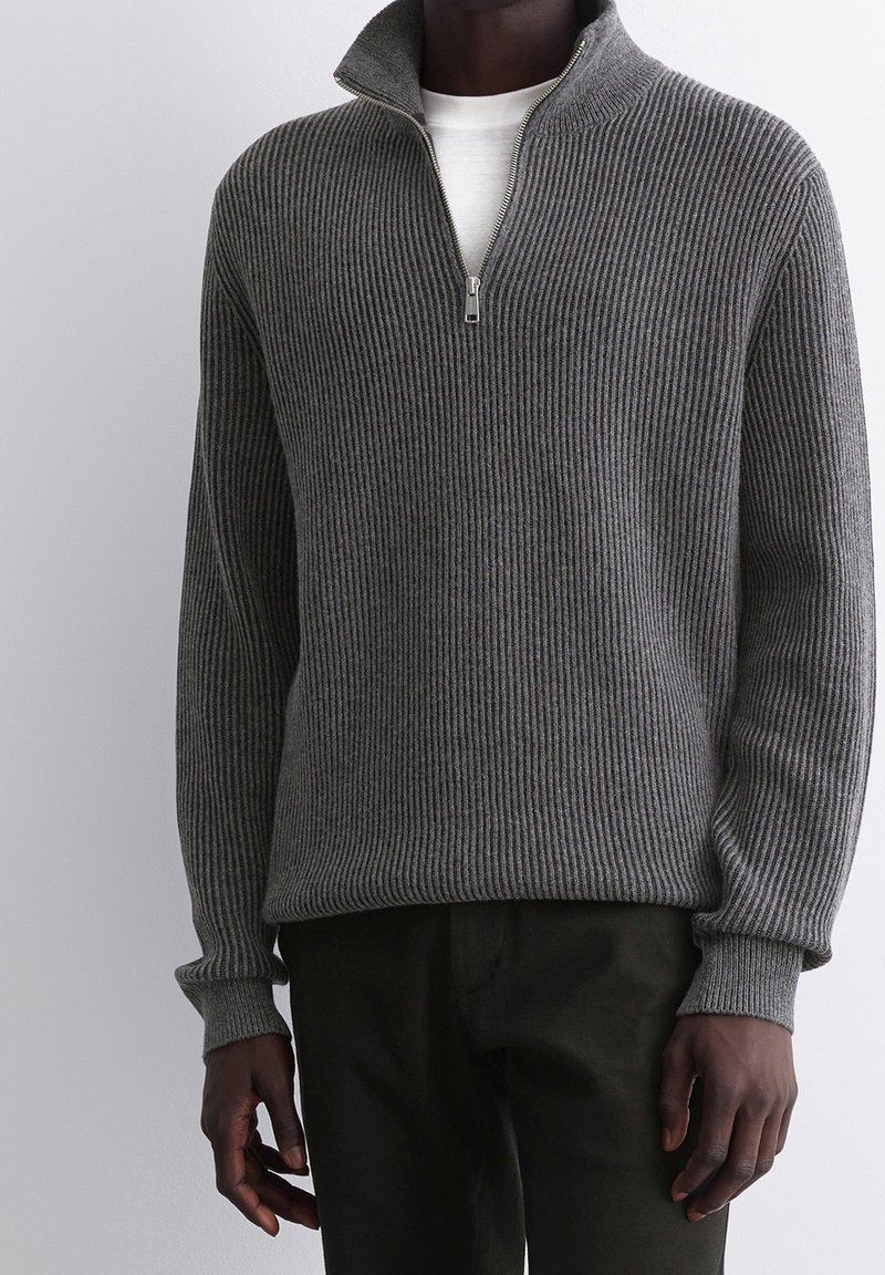Pull gris côtelé avec un col montant, doté d'une fermeture éclair demi-zip. Fabriqué en tissu tricot texturé, associé à un pantalon vert foncé.