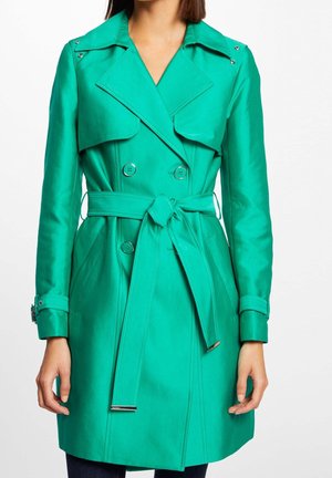 Femme portant un trench-coat double boutonnage bleu sarcelle vif avec ceinture nouée à la taille et poignets boutonnés, debout sur fond blanc.