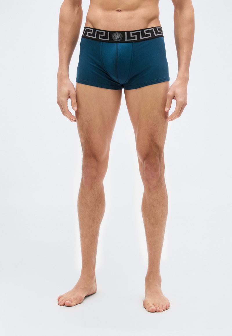 Αρσενικός κορμός και πόδια που φορ ούν βερμούδες τύπου μπόξερ σε τυρκουάζ με μαύρη μέση που φέρει γεωμετρικό μοτίβο και κεντρικό έμβλημα, στέκονται ξυπόλητοι σε λευκό φόντο.