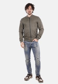 Giacca bomber verde oliva in tessuto morbido con zip frontale, tasche laterali, indossata con jeans chiari strappati e scarpe da ginnastica marroni.