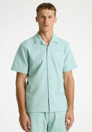Homme aux cheveux courts et bouclés portant une chemise à manches courtes boutonnée texturée vert clair et un pantalon assorti, debout devant un fond blanc.
