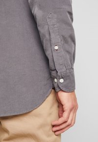 Manga de camisa de pana gris con botones, que presenta una sutil textura acanalada. El puño es redondeado y se puede ver un tejido en color beige por debajo.