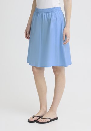 Kvinde iført en knælange lyseblå nederdel med elastisk talje, hvid top, sølvringe og sorte flip-flops stående på en hvid baggrund.