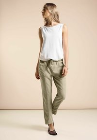 Top blanco sin mangas con cuello redondo, combinado con pantalones tapered de un verde oliva claro. La mujer lleva zapatos planos de malla negros. Diseño simple.