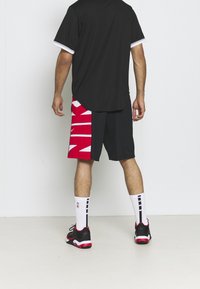 Maillot de sport noir avec des accents blancs, associé à un short rouge et noir arborant le bold texte blanc "NIKE". Chaussettes blanches et baskets noires.