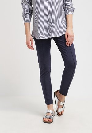 Personne portant un pantalon slim bleu marine, une chemise à rayures verticales à manches longues et des sandales à boucle argentée, debout sur un fond uni clair.