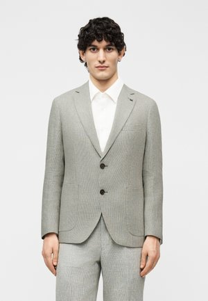 Mężczyzna o kręconych czarnych włosach, ubrany w jasnoszary garnitur na dwa guziki i białą koszulę, stojący na jednolitym jasnym tle.