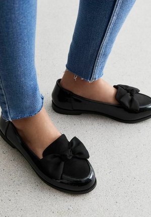 Fødder iført sorte laklæder loafers med store sløjfer, parret med blå denim jeans med frayed kant på et lyst, plettet gulv.