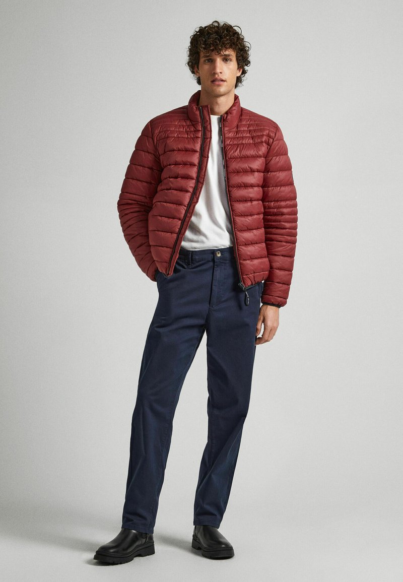 Młody mężczyzna w bordowej kurtce puchowej, białej koszulce, granatowych spodniach i czarnych butach, stoi na tle jednolitego, jasnoszarego tła.