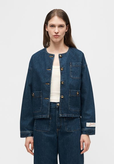 Γυναίκα που φοράει σκούρο μπλε τζιν μπουφάν με μεγάλα κουμπιά και ταιριαστό ψηλόμεσο τζιν παντελόνι, στέκεται μπροστά σε απλό φόντο.